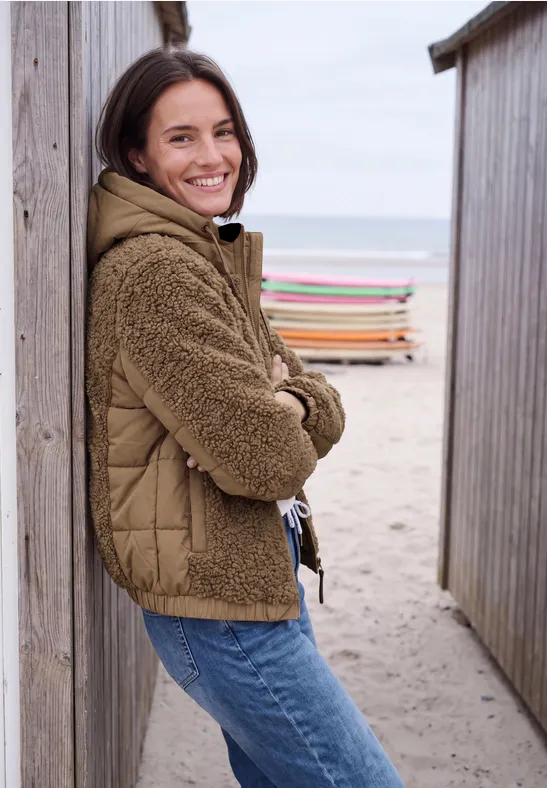 Une jeune femme souriante appuyée entre des cabanes de plage au bord de la mer.