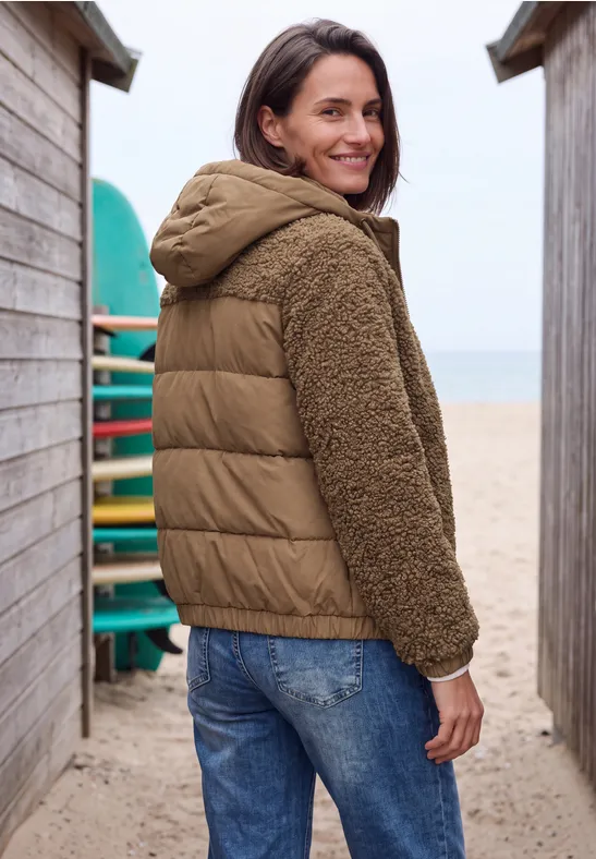 Femme en veste matelassée et polaire marron entre deux cabanes de plage, souriante, regard en arrière.