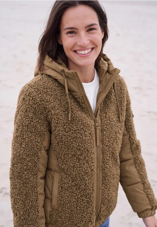 Femme souriante sur la plage, portant une veste polaire marron à capuche.