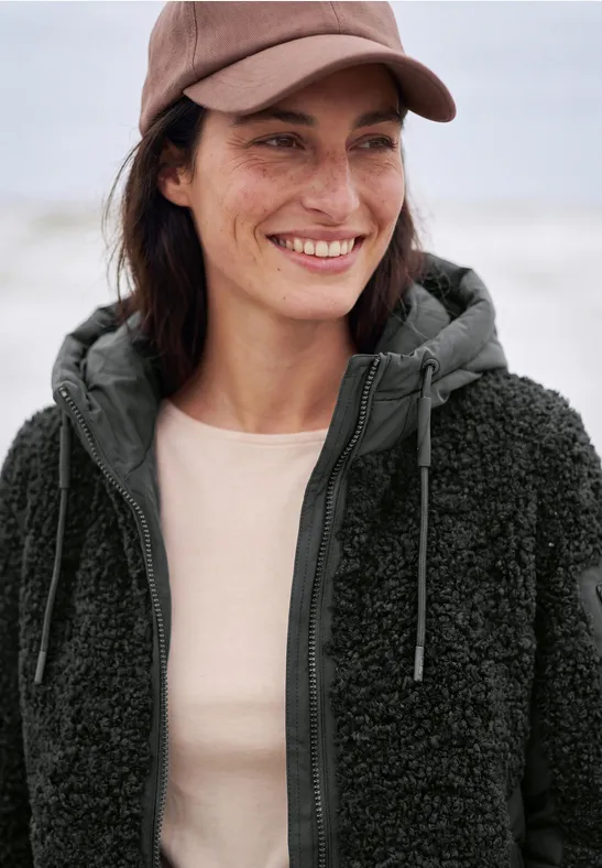 Une femme souriante en casquette et veste polaire au bord de la mer.
