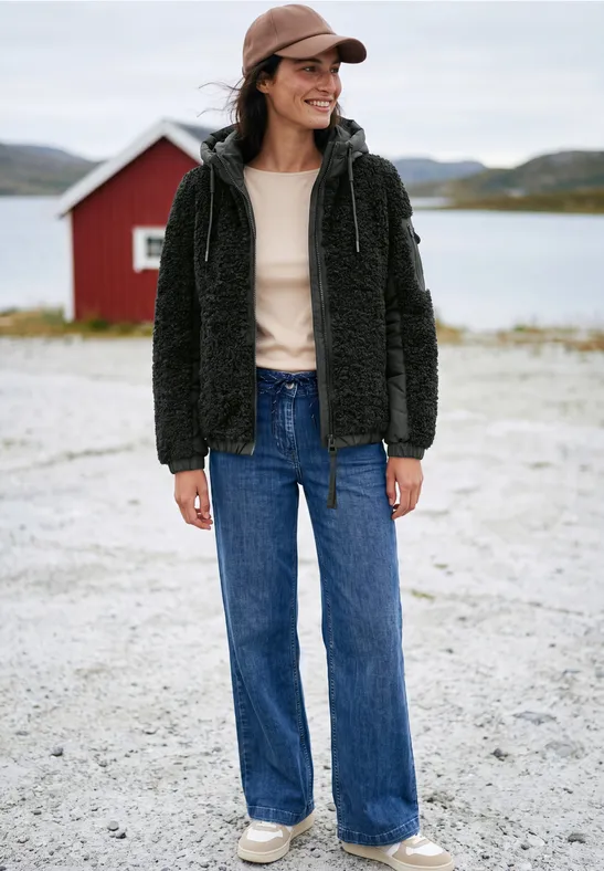 Femme en veste noire en fourrure, jean large et casquette, debout devant une cabane rouge au bord d’un lac.