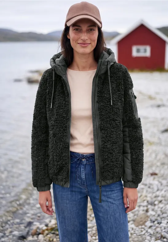 Femme en veste polaire noire et jean debout sur un rivage caillouteux devant une maison rouge.