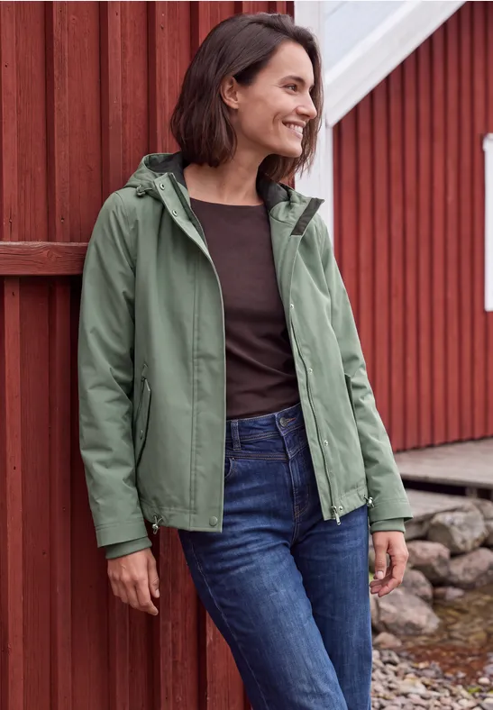 Een vrouw staat voor een rood houten gebouw, draagt een groene capuchonjas en jeans, en lacht opzij.