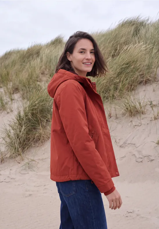 Frau in roter Jacke steht auf Sanddüne, schaut lächelnd über die Schulter, Dünen und Gras im Hintergrund.