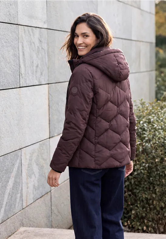 Frau in dunkelroter Steppjacke mit Kapuze steht draußen an einer Mauer und schaut lächelnd zurück.