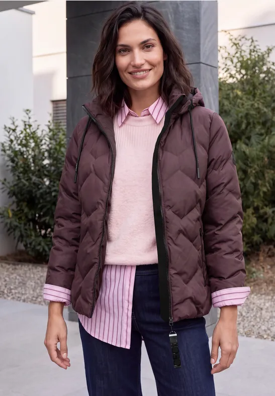 Frau steht lächelnd vor einem Haus mit Sträuchern, trägt eine braune Steppjacke und rosa-gestreiftes Hemd.