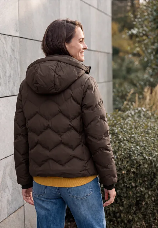 Person in brauner Daunenjacke und Jeans, lächelnd vor grauer Wand und Sträuchern.