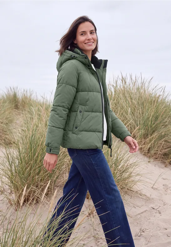 Frau in grüner Daunenjacke spaziert am windigen Strand mit Dünen im Hintergrund.