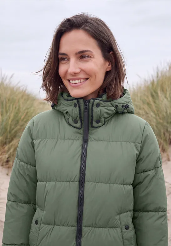Eine lächelnde Frau in einer grünen Steppjacke mit Kapuze, steht am Strand.