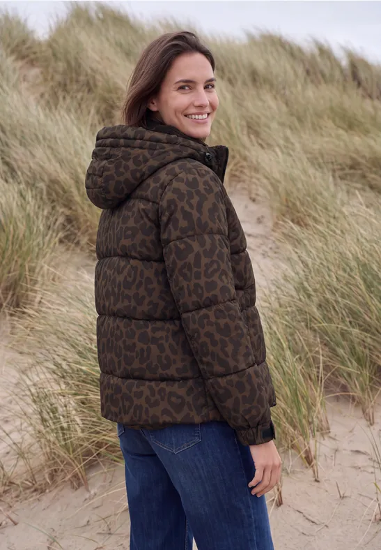 Frau in brauner, gemusterter Daunenjacke steht lächelnd in Dünenlandschaft.