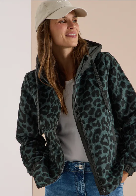 Frau in grüner Leo-Musterjacke, weißem Shirt und Jeans, Blick nach rechts, lächelnd.