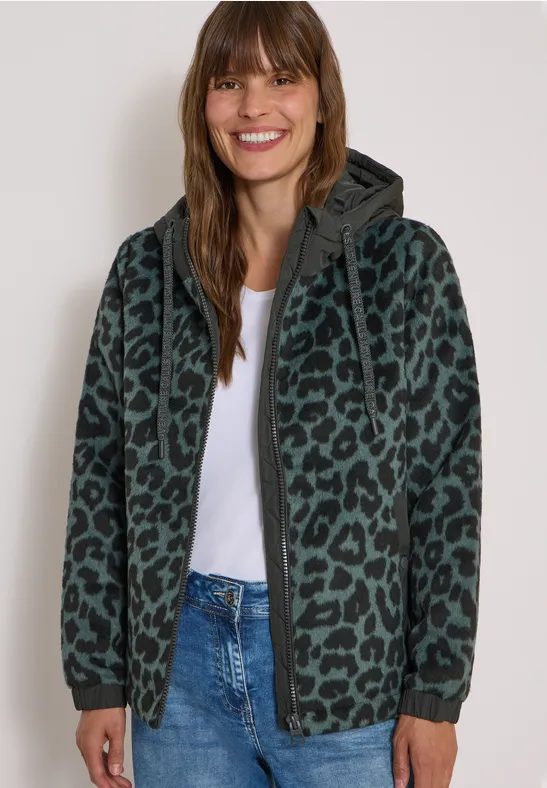 Lächelnde Frau in Jeans und weißem Shirt, trägt einen grünen Kapuzenpullover mit Leopardenmuster.