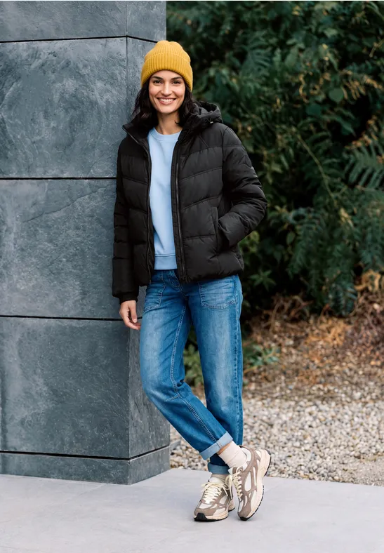 Eine junge Frau lehnt lächelnd an einer Wand, trägt schwarze Steppjacke, blaue Jeans, gelbe Mütze.