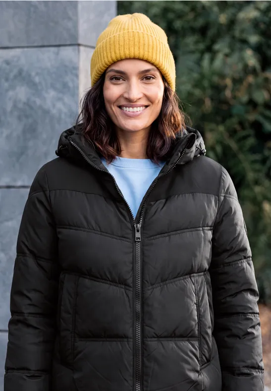 Eine lächelnde Person in einer schwarzen Steppjacke und gelber Mütze vor einer Steinwand.