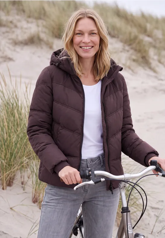 Een lachende vrouw in een bruine donsjas en spijkerbroek met haar fiets bij kustduinen.
