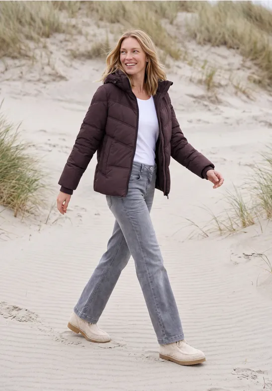 Een vrouw in een gewatteerd jack, spijkerbroek en sneakers wandelt glimlachend langs de duinen op het strand.