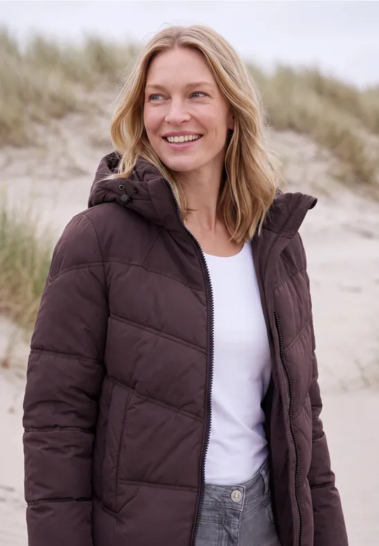 Een glimlachende vrouw in een bruine donzen jas en wit shirt op het strand voor zandduinen.