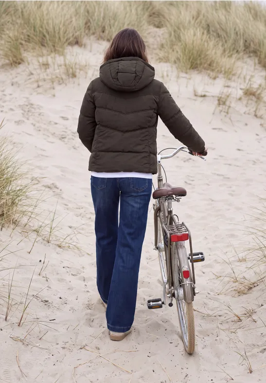 Person in Winterjacke und Jeans schiebt Fahrrad durch den Sand zwischen Dünen.