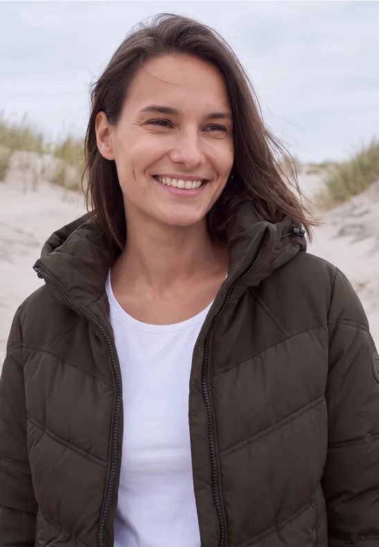 Frau mit Jacke und weißem T-Shirt lächelt am Strand vor Dünen.