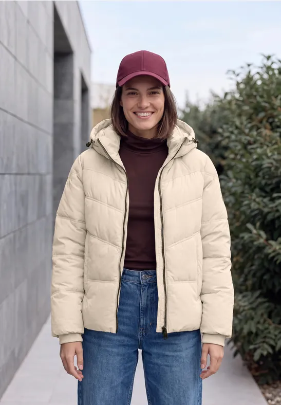 Junge Frau in beiger Daunenjacke und roter Mütze lächelt vor Hauswand und Pflanzen.
