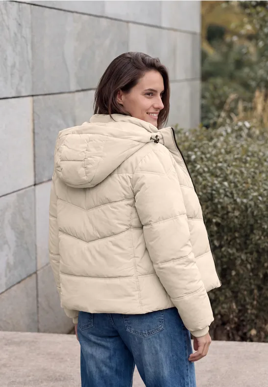 Frau in cremefarbener Steppjacke, lehnt an einer Steinwand und blickt lächelnd zurück.