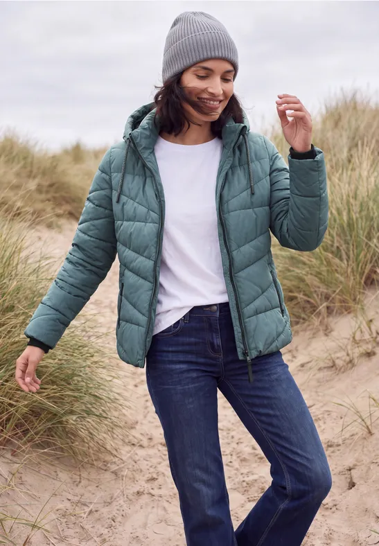 Eine Frau mit grauer Mütze, grüner Steppjacke, weißem T-Shirt und Jeans spaziert über Dünen am Strand.