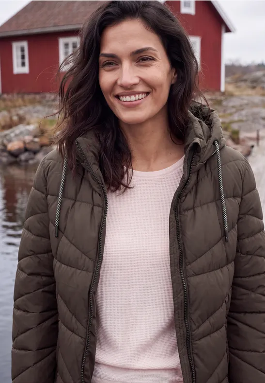 Lachende vrouw in een bruin winterjack en roze trui voor een rood huis aan het water.