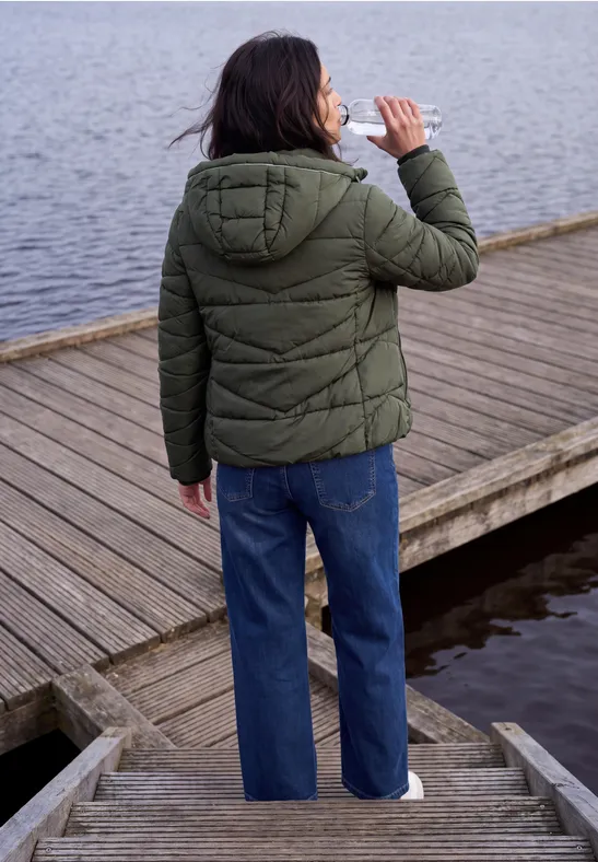 Person mit grüner Steppjacke und blauer Jeans trinkt am Holzsteg, Rückenansicht.