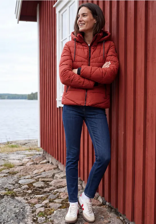 Frau lehnt in roter Steppjacke und Jeans entspannt an einem roten Holzhaus am Wasser.