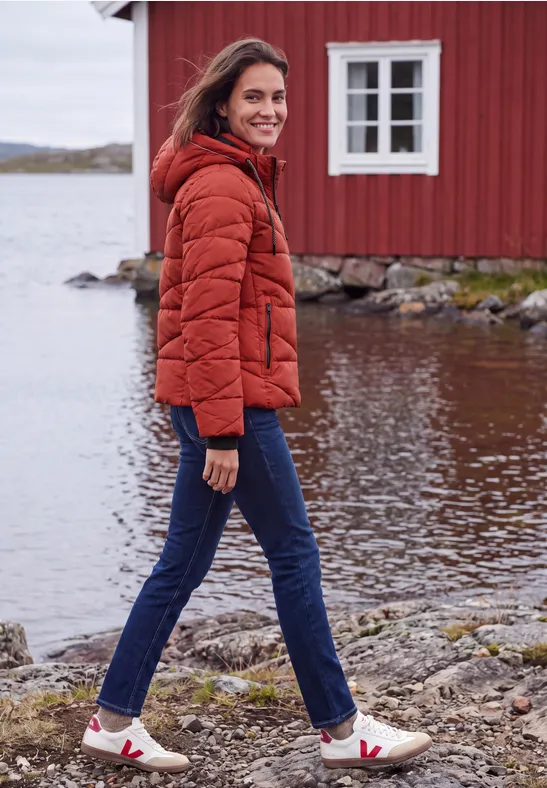 Eine Frau in oranger Steppjacke und Jeans läuft am Ufer vor einer roten Hütte am See.