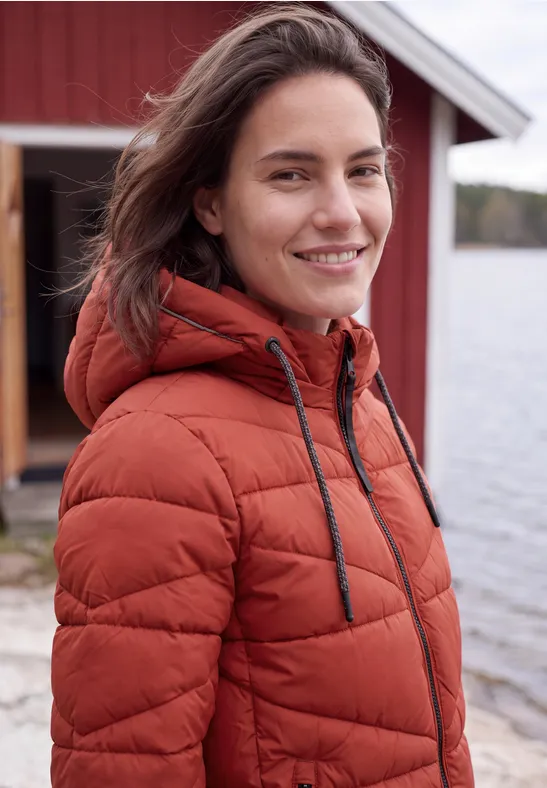 Lächelnde Frau in oranger Steppjacke vor rotem Holzhaus am See