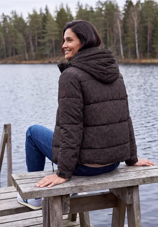 Vrouw zit glimlachend in donsjas op een houten bank bij een meer, omringd door bos en rustig water.