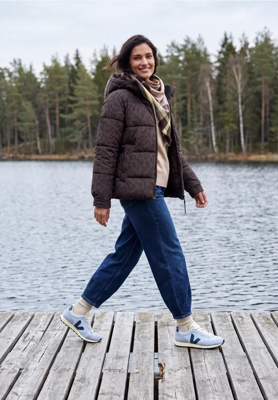 Vrouw in winterjas en sneakers loopt lachend over een steiger bij een meer.