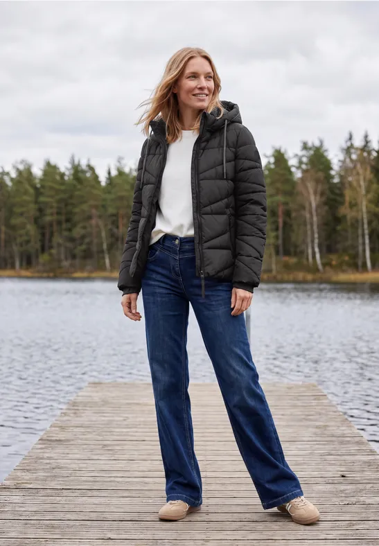 Frau in schwarzer Daunenjacke, weißem Pullover, blauen Jeans und Hausschuhen steht lächelnd auf einem Steg am See.