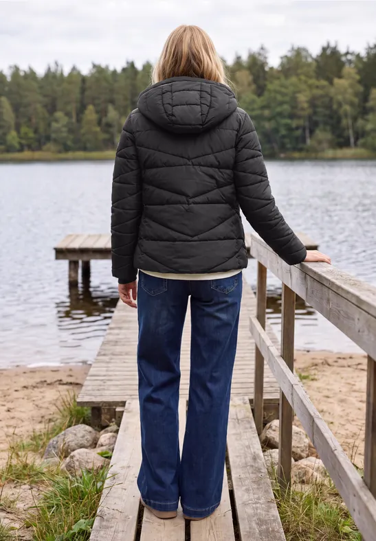 Eine Person mit schwarzer Steppjacke und Jeans steht auf einem Steg am ruhigen Waldsee.
