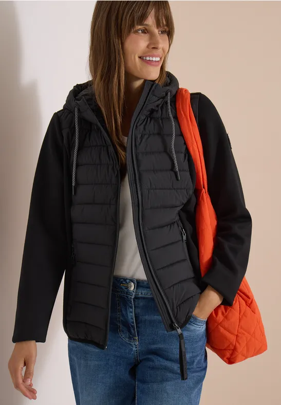 Femme en veste matelassée noire, jean et débardeur blanc, sac orange, sourire, fond neutre.