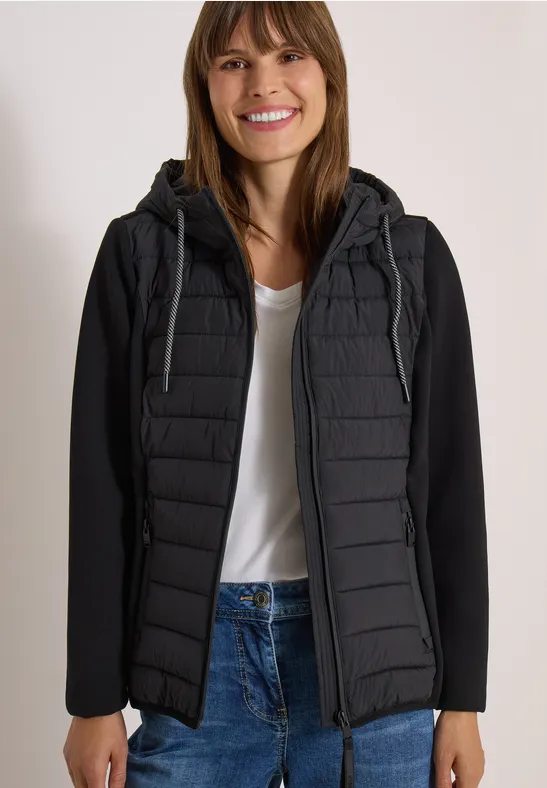 Une femme souriante porte une veste matelassée noire à capuche, un t-shirt blanc et un jean.