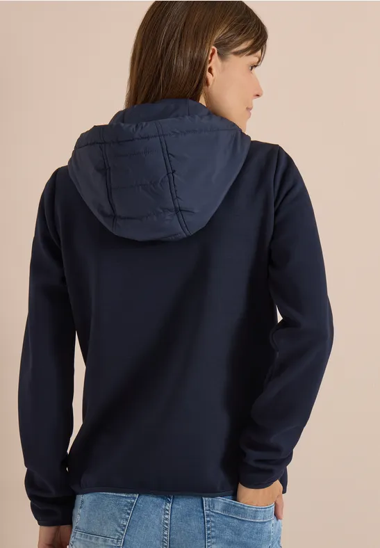 Frau von hinten in dunkelblauem Kapuzenpullover und Jeans vor hellbeigem Hintergrund.