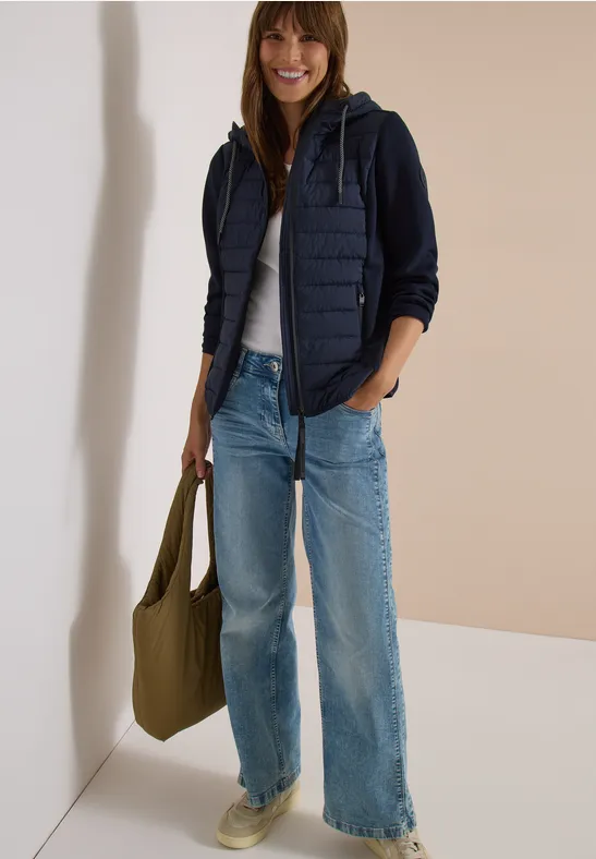 Lächelnde Frau trägt Jeans, weißes T-Shirt, dunkelblaue Kapuzenjacke mit Steppweste und hält eine grüne Tasche.