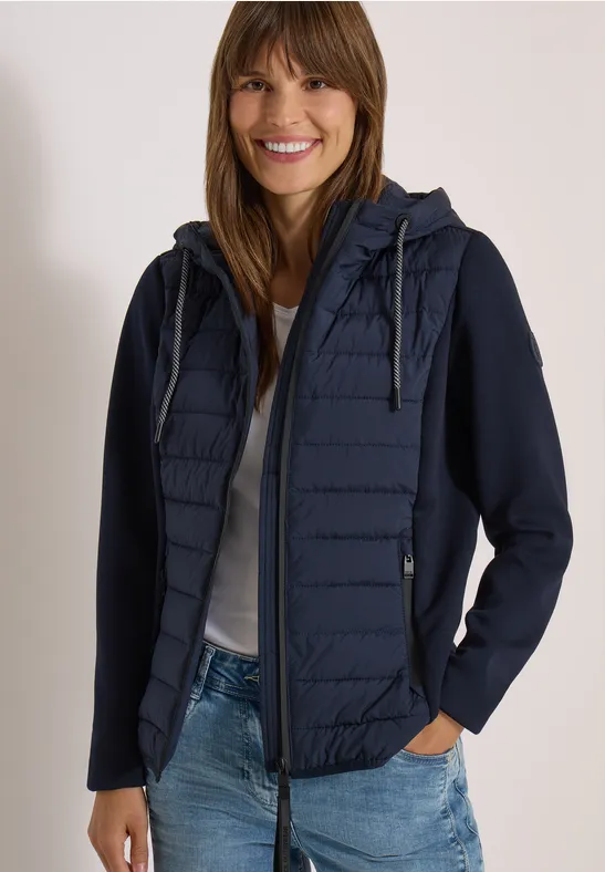 Eine lächelnde Frau in dunkelblauer, gesteppter Kapuzenjacke, weißem Shirt und Jeans.