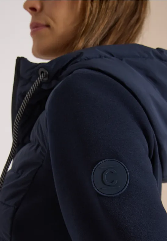 Frau in seitlicher Ansicht trägt eine dunkelblaue, gesteppte Kapuzenjacke mit rundem Logo-Patch am Ärmel.