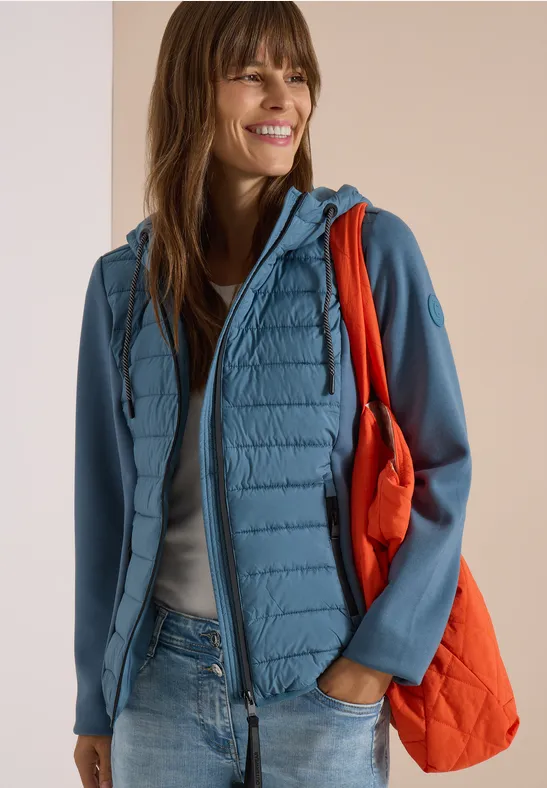 Lächelnde Frau in hellblauer Steppjacke mit Softshell-Ärmeln, Jeans und orangefarbener Tasche.