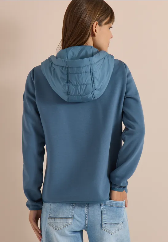 Frau in blauer Kapuzenjacke mit gesteppter Kapuze und Jeans, Rückansicht vor neutralem Hintergrund.