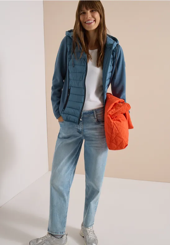 Frau in lässiger Jeans, weißem T-Shirt, grüner Steppjacke und orangefarbener Tasche, sie lächelt.