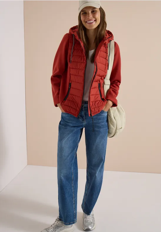 Frau mit roter Kapuzenjacke, roter Steppweste, blauen Jeans, weißen Sneakers und Cap vor beigem Hintergrund.