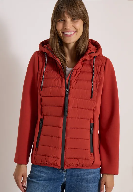 Eine Frau in roter gesteppter Kapuzenjacke und Jeans lächelt vor hellem Hintergrund.
