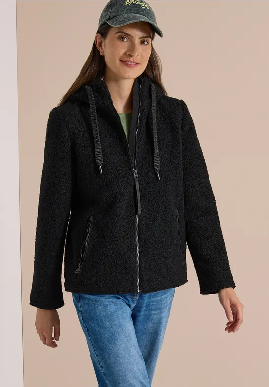 Frau trägt schwarze Teddyjacke mit Reißverschluss, Kapuze und Kordeln, kombiniert mit Jeans und Cap.