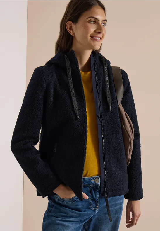 Eine lächelnde Frau trägt eine marineblaue Teddyjacke, gelben Pullover und Jeans, mit Schultertasche.
