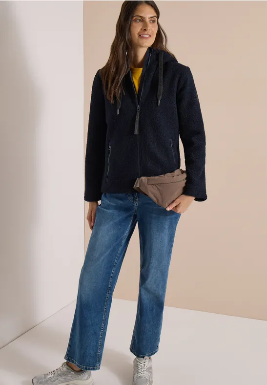 Frau in dunklem Wollmantel, Jeans und gelbem Pullover, hält eine braune Clutch vor hellem Hintergrund.