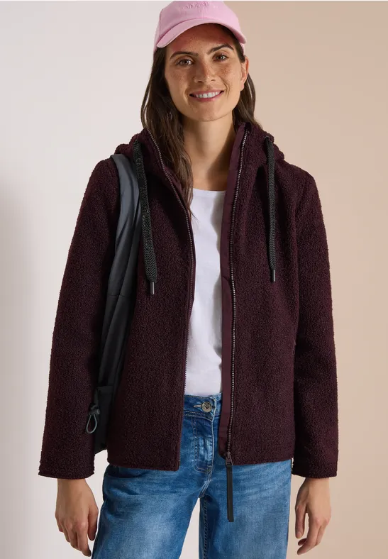 Frau in dunkelroter Teddy-Fleecejacke, weißem T-Shirt, Jeans und rosa Cap, mit Rucksack und Lächeln.